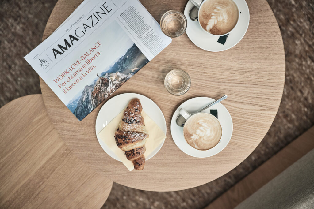 Hotelmagazin vom Workation Aparthotel AMA Stay auf einem Tisch mit Croissant und zwei Cappuccini